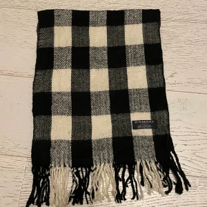 Rutig svartvit halsduk från Burberry - Snygg halsduk från Burberry i klassiskt rutigt mönster med svarta och vita rutor. Tillverkad i 100% kashmir för extra mjuk och lyxig känsla. Halsduken har fransar i ändarna och en diskret Burberry-label. Perfekt för att lyfta din vinterstil.