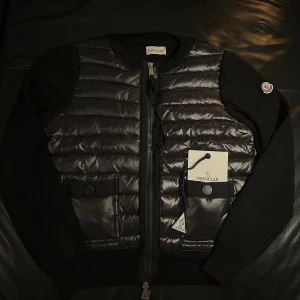  Moncler Cardigan  - Snygg svart jacka från Moncler med quiltad front och stickade ärmar. Jackan har rund hals, två fickor med knappstängning och dragkedja framtill. Moncler-logga på ärmen och diskreta detaljer som ger en exklusiv vibe.