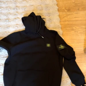 Svart hoodie från Stone Island - Svart hoodie från Stone Island med klassisk logopatch på ärmen och bröstet. Tröjan har huva med snörning och en stor magficka framtill. Mjuk insida och ribbade muddar vid ärmslut och nederkant. Perfekt för dig som gillar streetwear och snygga detaljer.