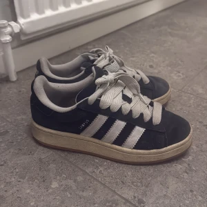 Adidas Campus sneakers svart/vit - Säljer ett par klassiska Adidas Campus sneakers i svart mocka med vita detaljer och de ikoniska tre ränderna på sidorna. Skorna har vit snörning, platt sula i gummi och rund tå. Perfekta för dig som gillar retrostil och streetwear.