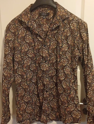 Slim fit blommig skjorta från SMOG - Slim fit skjorta från SMOG med blommigt mönster i brunt, beige, vitt och inslag av grönt. Skjortan har klassisk krage, långa ärmar och knappar framtill. Materialet är mjukt och känns som bomull, perfekt för dig som gillar detaljerade prints och unik stil.