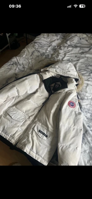 Vit dunjacka från Canada Goose - Säljer en vit dunjacka från Canada Goose med huva. Jackan har två stora fickor framtill med lock och dragkedja, samt klassisk Canada Goose-logga på ärmen. Perfekt för kalla vinterdagar och har en snygg, stilren look.