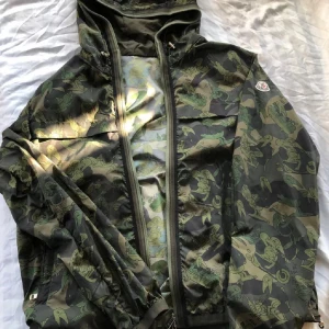 Moncler camo vindjacka med mönster - Säljer en grön och mörkgrön vindjacka från Moncler med unikt camouflage-mönster och tecknade figurer. Jackan har huva, hel dragkedja. 