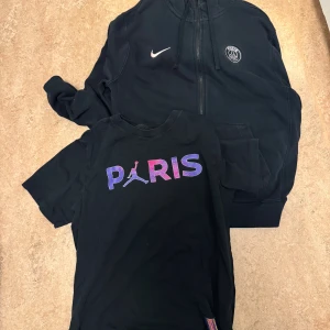 Svart Nike PSG hoodie med dragkedja - Svart hoodie från Nike x Paris Saint-Germain med PSG-logga på bröstet och vit Nike swoosh. Jackan har huva med snörning, två fickor fram och hel dragkedja. Perfekt för dig som gillar sportig streetstyle och vill representera Paris.