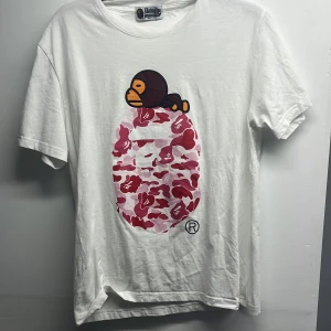 Vit BAPE t-shirt med rosa camo - Vit t-shirt från A Bathing Ape med ikoniskt BAPE-tryck i rosa och röd camo på bröstet samt en liten apa ovanpå. Klassisk rund hals och korta ärmar. Liten BAPE-logga på ärmslutet. Perfekt för dig som gillar streetwear och vill sticka ut.