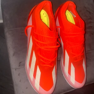 Adidas Crazyfast elite röda fotbollsskor ag - Säljer ett par Adidas Crazyfast fotbollsskor i en riktigt snygg röd färg med vita ränder på sidorna. Skorna har snörning och en rund tå, samt en lätt och ventilerande ovandel i syntetmaterial. Perfekt för dig som vill sticka ut på planen.
