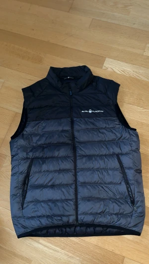 Svart dunväst från Sail Racing - Svart dunväst från Sail Racing med quiltad design och hög krage. Västen har två sidofickor med dragkedja och logga på bröstet. Materialet är lätt och syntetiskt, perfekt för lager-på-lager. Snygg och sportig look med normal passform.