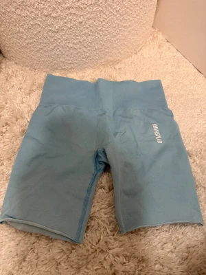 Ljusblå Gymshark biker shorts - Säljer ett par ljusblå biker shorts från Gymshark med hög midja och ribbad linning. Men jag har klippt dom då de var långa tights innan. Gymshark-loggan är tryckt på ena benet.