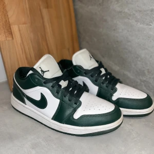 Nike Air Jordan 1 Low vit/grön - Säljer ett par Nike Air Jordan 1 Low i vitt och mörkgrönt skinn med klassisk svart swoosh och broderad Air Jordan-logga på hälen. Säljes då de är för stora för mig, svårt att hitta just dessa gröna! Antäva fåtal gånger, kanske 5 max!!🍀💚