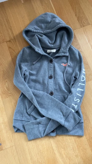 Grå hoodie från Hollister med knappar - Grå hoodie från Hollister i mjuk bomull med knappar framtill och huva. Långärmad modell med logotyp i orange på bröstet och tryck på ena ärmen. Perfekt för chill dagar och enkel att matcha med jeans eller joggers.