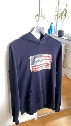 Mörkblå hoodie från Ralph Lauren - Snygg mörkblå hoodie från Ralph Lauren Denim & Supply med stor amerikansk flagga-patch och broderade detaljer på bröstet. Tröjan har huva, magficka och är tillverkad i 100% bomull. Perfekt för dig som gillar ”archive” stilen!! Storleken är M.