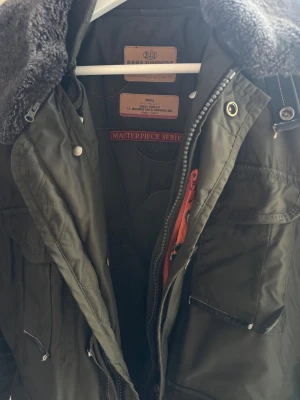 Grön parkajacka från Parajumpers - Säljer en mörkgrön parkajacka från Parajumpers, flera fickor och snygga broderade detaljer på ärmen. Jackan har dragkedja, knappar och meshficka på insidan. Perfekt för kalla dagar och riktigt snygg streetstil. Jackan är använd så små skavanker finns men inget som påverkar funktionen.