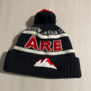 Mörkblå Åre mössa - Säljer en mörkblå stickad mössa med vit-röda detaljer och tofs i rött och mörkblått. Mössan har bred uppvikt kant, broderat bergmotiv och texten 'ÅRE' framtill. 