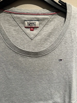 Grå t-shirt Tommy hilfiger   - Snygg grå t-shirt från Tommy Jeans i storlek XS. Klassisk rund halsringning och korta ärmar. Liten broderad logga på bröstet och diskreta röda och blå sömmar längs insidan av halsen och sidan. 