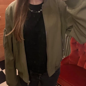 Olivgrön bomberjacka med dragkedja - Snygg olivgrön bomberjacka med klassisk ribbad mudd vid ärmslut och nederkant. Jackan har en silverfärgad dragkedja framtill och en extra ficka med dragkedja på ärmen. Materialet är slätt och känns lätt, perfekt för dig som gillar streetstyle.