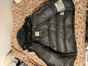 Svart Moncler pufferjacka med huva - Svart pufferjacka från Moncler med huva och flera dragkedjeförsedda fickor. Jackan har ett quiltat mönster och är fodrad med dun för extra värme. Insidan har en unik serietidningsetikett. Perfekt för kalla dagar och snygg streetstil.