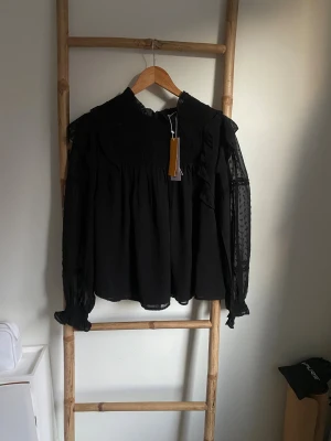 Svart boho blus med spetsdetaljer - Svart blus från Y.A.S med boho-vibbar, volangdetaljer och transparenta långärmade partier med små prickar. Spetsdetaljer och smock upptill. Perfekt för dig som gillar att sticka ut med stil.