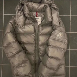 Grå Moncler pufferjacka med huva - Säljer en grå pufferjacka från Moncler med huva och klassisk logga på ärmen. Jackan har dragkedja framtill, praktisk ficka på ärmen och quiltad design. Perfekt för kalla dagar och riktig snygg! Den är använd en säsong så finns ju skavanker på knappar oså men inte några hål eller liknande! 