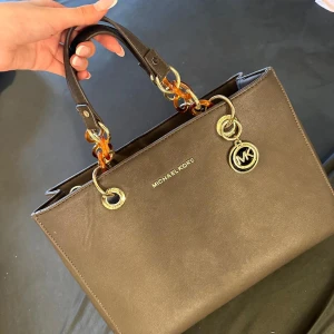 Mk väska grå nästan nya  - Snygg brun handväska från Michael Kors med guldfärgade detaljer och MK-logga. Väskan har rejäla handtag med guldfärgade 