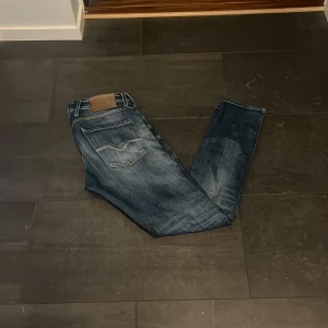 Replay Anbass jeans blå straight fit - Snygga Replay Anbass jeans i blå tvätt med slitningar och klassiska fem fickor. Jeansen har rak passform, normal midja och detaljerade sömmar på bakfickorna. Materialet är robust denim med en cool faded effekt. Perfekt för dig som gillar stilrena och trendiga jeans.
