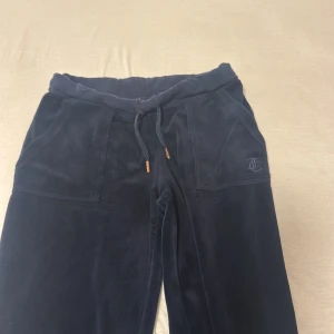 Mörkblå mjukisbyxor Juicy Couture XS - Mörkblå mjukisbyxor från Juicy Couture i storlek XS. Byxorna har snörning i midjan, två stora fickor fram och bak med silverfärgade knappar, samt broderad logga på ena fickan. Materialet är mjukt och lyxigt, perfekt för chill dagar.