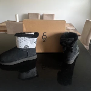 Svarta UGG boots med rosett - Säljer ett par svarta UGG boots med fluffig päls runt kanten och en snygg rosett bak på hälen. Skorna har rund tå, platt sula och är tillverkade i mjuk mocka med UGG-logga bak. Perfekta för kalla dagar och har en klassisk siluett.