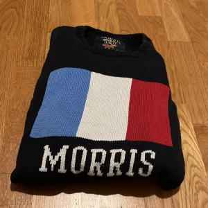 Svart stickad tröja Morris M/L - Svart stickad tröja från Morris i storlek Large med fransk flagga i blått, vitt och rött på bröstet samt Morris-logga under. Rund hals och ribbade muddar vid ärmslut och nederkant. Snygg detalj med ett litet M i guld på ena ärmen. Storleken passar både M och L, skicket är mycket fint! Hör av dig om du har några frågor