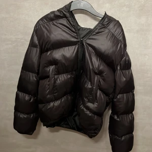 Svart pufferjacka med huva - Säljer en svart pufferjacka med huva och dragkedja framtill. Jackan har en snygg, lite glansig yta och är vadderad i breda sektioner för extra värme. Passformen är normal och jackan har långa ärmar. Perfekt för kalla dagar. 