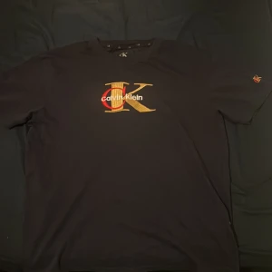Svart Calvin Klein t-shirt med broderi - Svart t-shirt från Calvin Klein i 100% bomull, med stort broderat CK-logo i guld och rött på bröstet samt mindre broderi på ärmen. Klassisk rund hals och korta ärmar. Perfekt för dig som gillar stilrena märkesplagg med snygga detaljer.