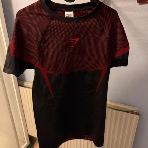 Gymshark Onyx V5 red M - Snygg svart och röd t-shirt från Gymshark med sportig design och korta ärmar. Materialet är lätt och andas, perfekt för träning. T-shirten har ett mönstrat parti upptill och Gymshark-logga på bröstet. Passar dig som gillar att sticka ut på gymmet.