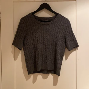 Grå stickad tröja från C, kortärmad - Säljer en grå stickad tröja från Cubus i storlek XS. Tröjan har ett snyggt kabelstickat mönster, rund hals och korta ärmar. Perfekt för dig som gillar klassisk stil med en modern twist. Passar dig som vill ha något bekvämt och stilrent.