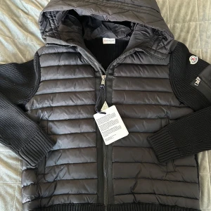 Svart Moncler cardigan med huva - Svart cardigan från Moncler med stickade ärmar och huva. Jackan har dragkedja fram, ribbade muddar och en ficka med dragkedja på ärmen. Ikonisk Moncler-logga på vänster arm. Perfekt för dig som vill ha en clean och stilren look. Passar bäst L!