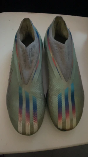 Adidas  fotbollsskor elite elite med färgglada ränder - Säljer ett par Adidas fotbollsskor elite elite  i ljusgrön och grå färg med tre ikoniska ränder i blått, vitt och rosa på ovansidan. Skorna har en strömlinjeformad design utan snörning, mönstrad ovandel och dobbar för gräsplan. Perfekt för dig som vill sticka ut på planen.