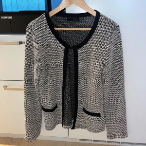 Svartvit kofta från Olivia - Snygg svartvit kofta från Olivia med långärmad design och två dekorativa fickor framtill. Koftan har ett klassiskt rutigt mönster och svarta kanter längs öppningen och fickorna. Perfekt för dig som gillar stilrena och tidlösa plagg.