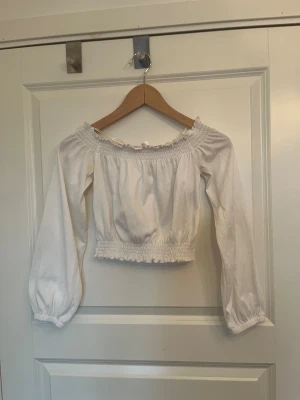 Vit croppad blus med offshoulder - Supersöt vit blus från H&M i storlek XS. Croppad modell med offshoulder och långa puffärmar. Smockad kant upptill och nedtill, samt volangdetaljer och knytband vid halsen. Perfekt för dig som gillar romantisk och trendig stil.