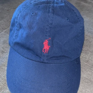 Mörkblå keps från Polo Ralph Lauren - Snygg mörkblå keps från Polo Ralph Lauren med klassisk röd broderad logga framtill och 'POLO' broderat baktill. Justerbar rem med metallspänne. Tillverkad i 100% bomull för skön passform och stilren look.