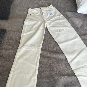 Vita wide jeans från Never Denim - Säljer ett par vita jeans från Never Denim med wide fit och hög midja. Byxorna har slitna detaljer på båda benen och klassisk femficksdesign. Materialet är jeans och modellen är rak och loose med regular wide passform. Perfekt för dig som gillar trendiga och avslappnade byxor.