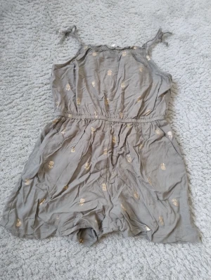 Grön romper med ananasmönster  - Supersöt grön kort romper/one-piece från H&M med tunna axelband som knyts och elastisk midja. Klänningen har små guldiga ananasmönster över hela tyget och är gjord i mjuk bomull. 