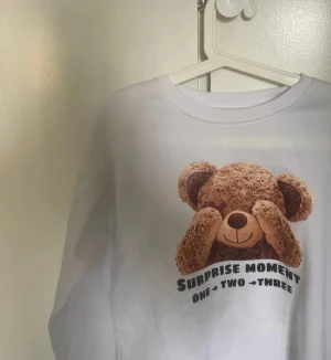 Vit sweatshirt med nalletryck - Vit sweatshirt med ett gulligt tryck av en nalle som håller tassarna för ögonen och texten 'Surprise Moment One Two Three' under. Tröjan har rund hals och långa ärmar, perfekt för dig som gillar söta och lekfulla motiv.ALDRIG ANVÄNT!