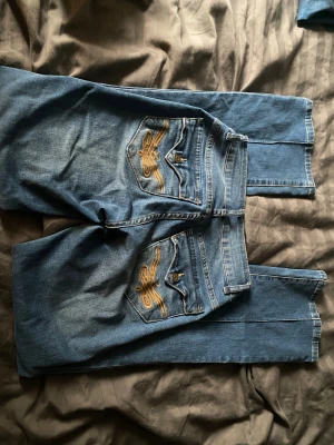Blå wide jeans från Madlady - Säljer ett par blå jeans från Madlady med bootcut och låg midja. Jeansen har dekorativa sömmar framtill, fickor med knappstängning bak och coola broderade detaljer i gult på bakfickorna. 32 Regular men sitter som xs. Varsamt använda 