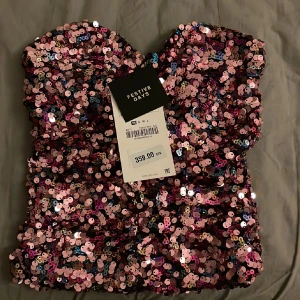 Glittrig paljett tubtopp från Zara - Säljer en glittrig tubtopp från zara Festive Days, täckt med rosa, lila och silverfärgade paljetter. Toppen är axelbandslös och har en croppad passform som ger en snygg siluett. Perfekt för att sticka ut med extra bling och färg.