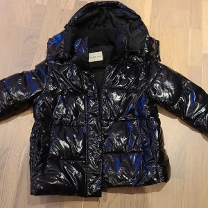 Glansig pufferjacka från Calvin Klein Jeans - Snygg svart jacka från Calvin Klein Jeans med glansig finish och huva. Jackan har dragkedja framtill, diskret CK-logga på bröstet och på en arm. Insidan har stor Calvin Klein Jeans-tryck. Perfekt för kalla dagar och riktigt stilren look.