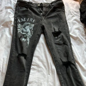Svarta skinny jeans från Amiri - Snygga svarta jeans från Amiri med skinny passform och slitna detaljer på benen. Framsidan har en stor vit ängelprint och AMIRI-logga. Klassisk femficksdesign, dragkedja och knapp med Amiri-märkning. Jeansen är gjorda i stretchig denim för skön känsla.