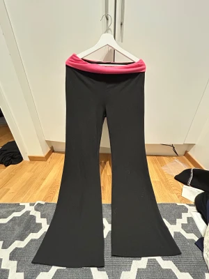 Svarta bootcut byxor med rosa midja - Snygga svarta bootcut byxor med bred rosa midja. Byxorna har en mjuk och stretchig känsla, perfekta för dig som gillar bekväma och trendiga plagg. Passformen är loose och midjan är låg, vilket ger en avslappnad vibe.