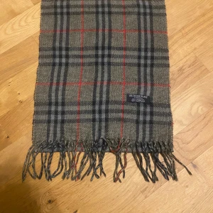 Burberry halsduk   - Snygg halsduk från Burberry i 100% kashmir med klassiskt rutigt mönster i beige, svart, grått och röda linjer. Mjukt material och fransar i ändarna. Perfekt för att lyfta din stil med en ikonisk accessoar från London.