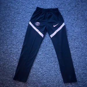 Svarta PSG träningsbyxor från Nike - Svarta träningsbyxor från Nike och PSG med rosa detaljer och loggor på båda sidor. Byxorna har en smal passform, elastisk midja och är gjorda i lätt polyester. Snygga rosa ränder går diagonalt över låren för extra stil.