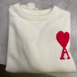 Vit stickad tröja från Ami Paris - Vit stickad tröja från Ami Paris med röd hjärtdetalj och bokstaven A på bröstet. Rund hals med ribbade muddar vid ärmslut och nederkant. Mjuk och skön kvalitet, perfekt för dig som gillar stilrena plagg med en twist.