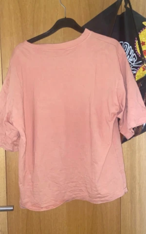 Rosa t-shirt med kattmotiv - T-shirt i en soft rosa färg med ett tryck av en katt i solglasögon och röd tröja med texten 'NO' på bröstet. Modellen är loose fit och har korta ärmar. Perfekt för dig som gillar roliga prints och chill vibe.