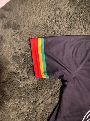 Ajax x Bob Marley t-shirt Adidas - Säljer en svart Ajax t-shirt från Adidas med Bob Marley-tema. T-shirten har korta ärmar med röd, gul och grön rand på mudden, samt tryck med citat, illustrationer och Ajax-logga. Materialet är polyester och modellen är normal passform.