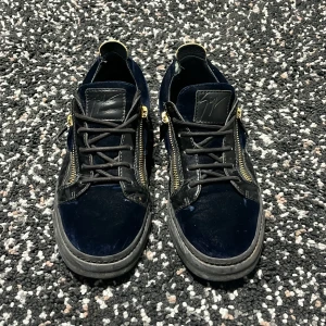 Giuseppe Zanotti blå sneakers med dragkedja - Säljer ett par mörkblå sneakers från Giuseppe Zanotti med coola guldfärgade dragkedjor på sidorna och snörning. Skorna har en mix av sammet och skinn, samt svarta detaljer och platt sula. Perfekt för dig som gillar lyxiga och unika sneakers.